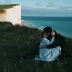 A couple shares a romantic embrace on the scenic cliffs of Seaford, UK, with a backdrop of the ocean and lighthouse.