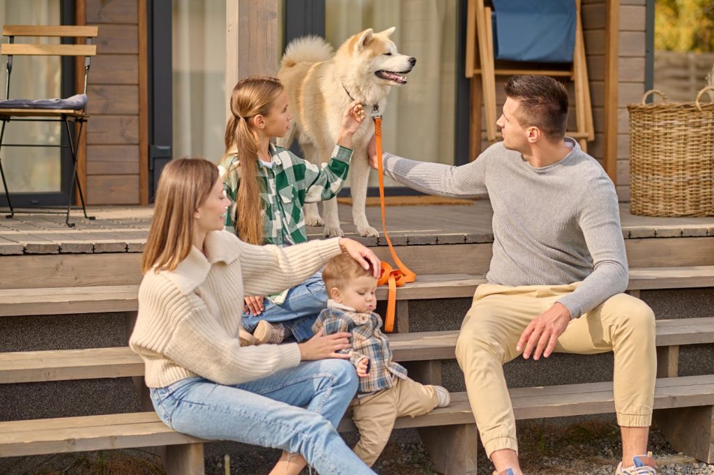 Family enjoying outdoor time on porch with dog, father holding leash, mother with toddler, and girl petting dog