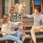 Family enjoying outdoor time on porch with dog, father holding leash, mother with toddler, and girl petting dog