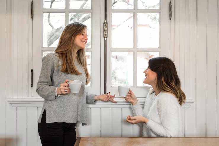 Two women stand by a window, each holding a coffee cup and enjoying a moment together