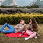 Couple enjoying a picnic with berries on a red blanket in a lush garden setting near a modern glass bridge