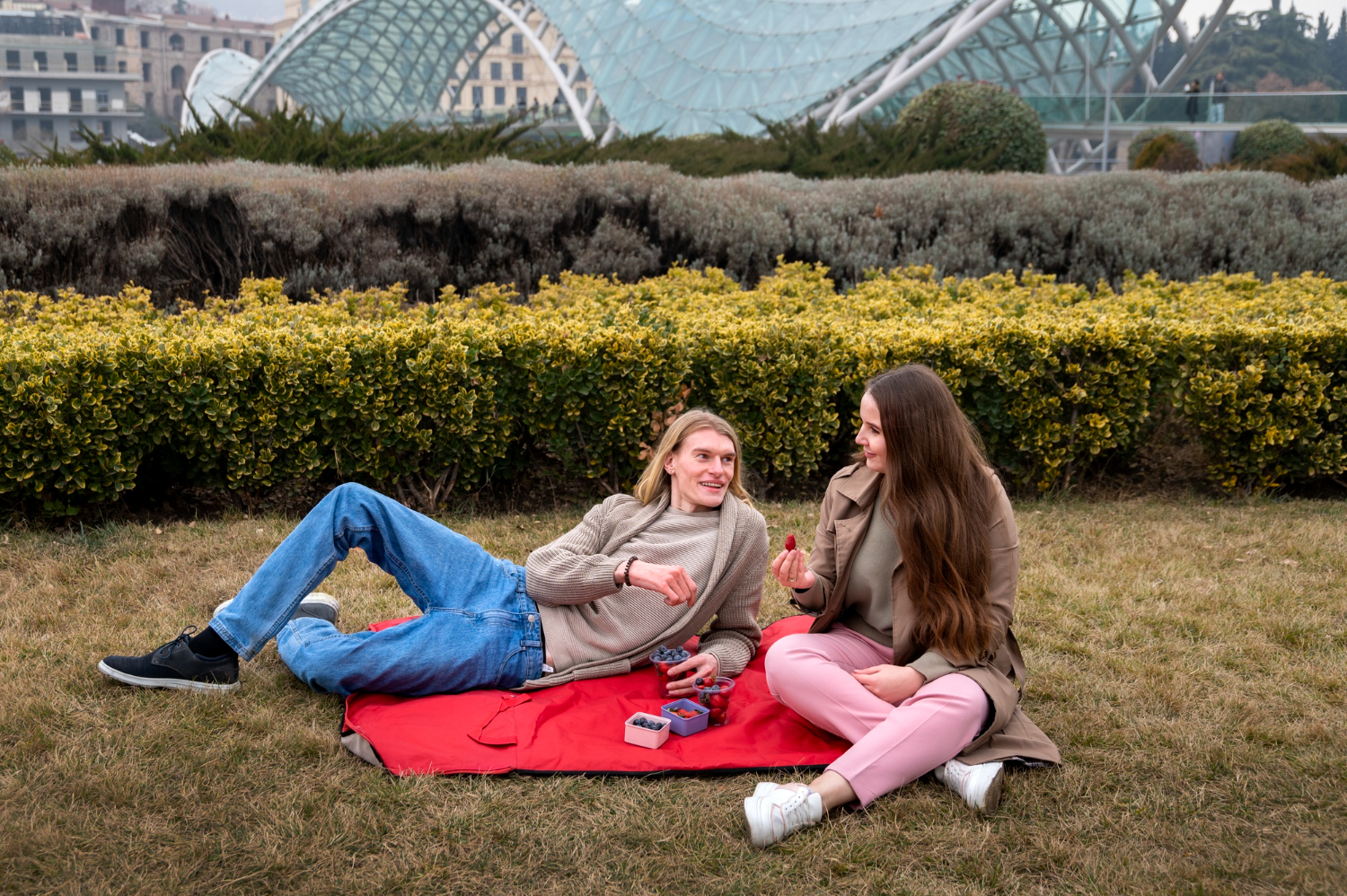 Couple enjoying a picnic with berries on a red blanket in a lush garden setting near a modern glass bridge
