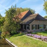 Charming cottage by a canal with a thatched roof, surrounded by lush greenery and vibrant flowers in a serene setting
