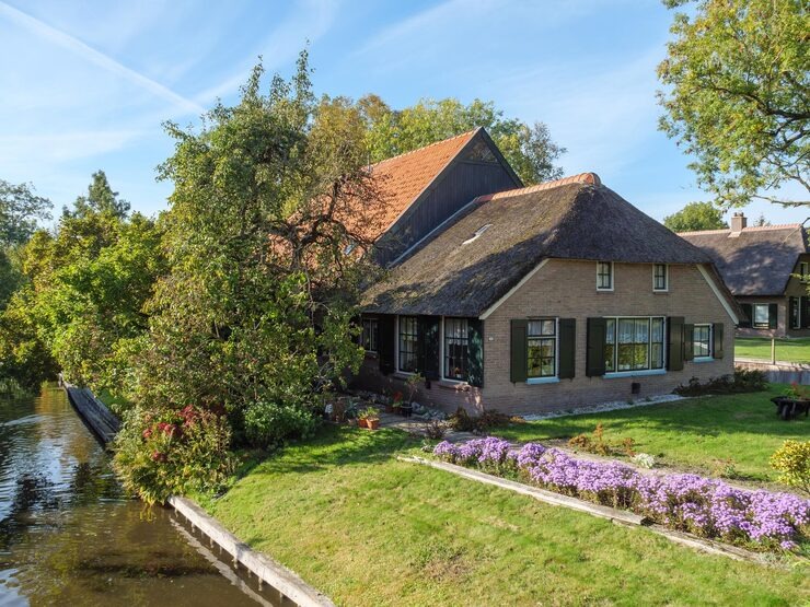 Charming cottage by a canal with a thatched roof, surrounded by lush greenery and vibrant flowers in a serene setting